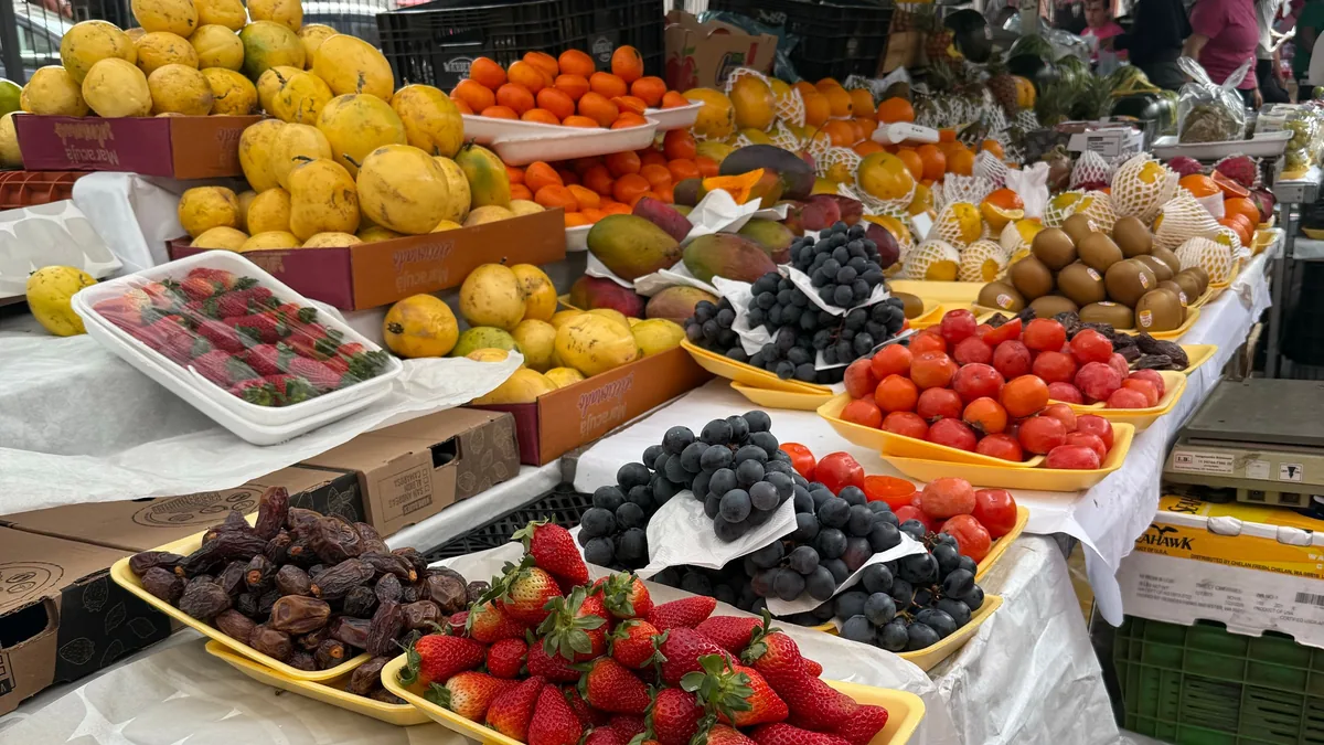 Banca de feira livre em São Paulo com bandejas de morango, cereja, uva, tomate e caixas de manga expostas sobre a mesa coberta de plástico branco