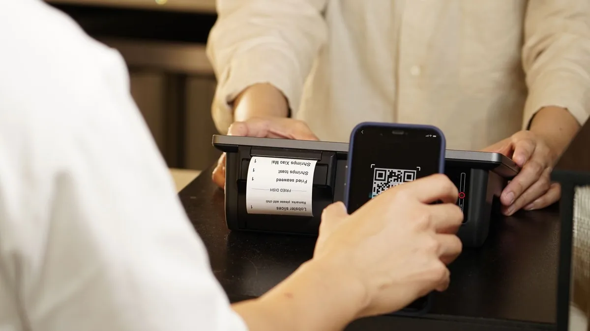 Mão aproximando um celular de uma maquininha de balcão para ler um QR Code, com a bobina de papel do pedido saindo ao lado — o mesmo gesto que está por trás do Pix Automático