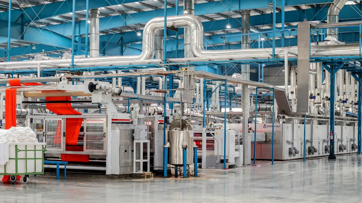 Galpão de fábrica têxtil com máquinas de produção industrial enfileiradas, tecido laranja saindo de um rolo e tubulações metálicas no teto