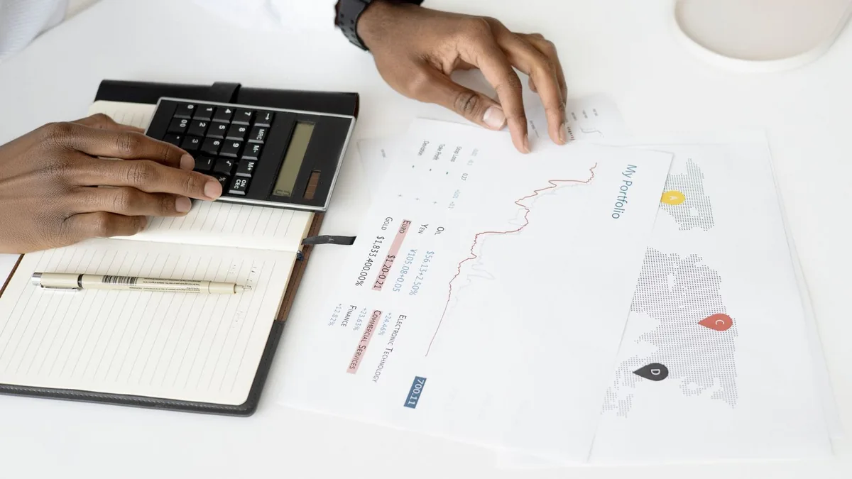 Vista de cima de uma mesa branca com uma pessoa segurando uma calculadora sobre um caderno aberto e conferindo um relatório impresso com gráfico de linha — o gesto de conferir contas que falta ao ROI de IA na maioria das empresas