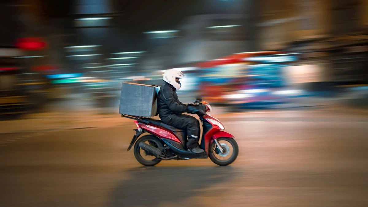 Entregador de capacete branco pilotando uma moto vermelha com baú cinza à noite, com as luzes da rua borradas pelo movimento